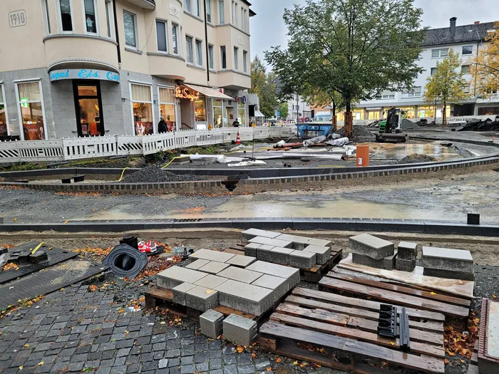 Baustelle in der Innenstadt mit auf Paletten gelagerten Pflastersteinen und Wasserlachen vor städtischen Gebäuden.
