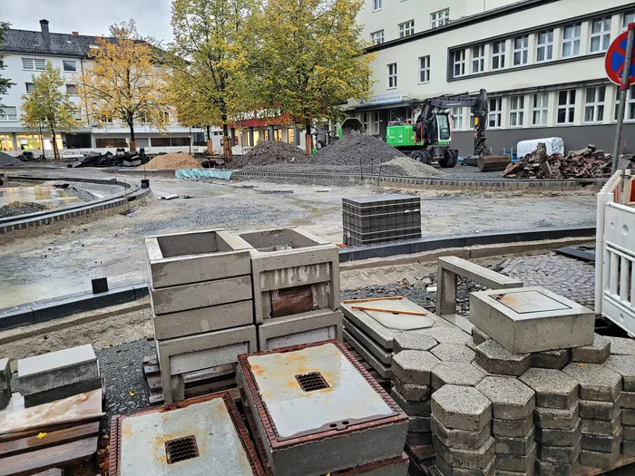 Baustelle in der Innenstadt mit Betonbauteilen, Pflastersteinen und einem Bagger vor städtischen Gebäuden und Bäumen.