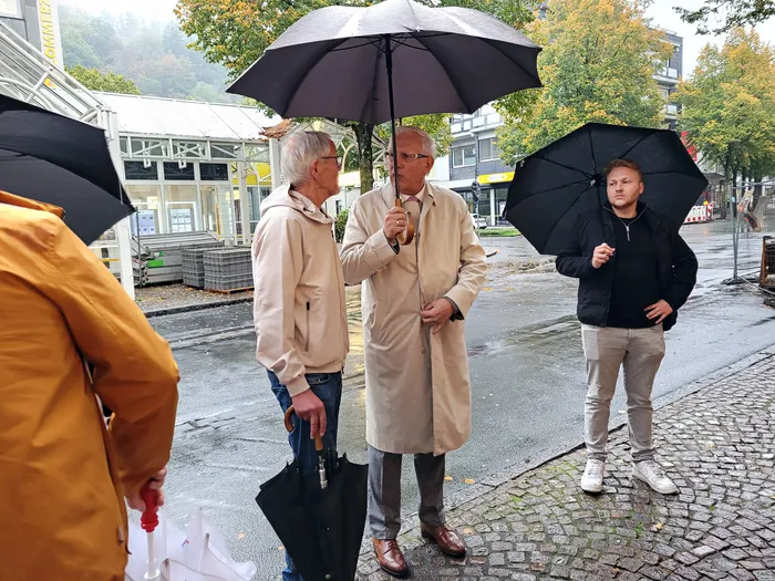 Drei Männer stehen unter schwarzen Regenschirmen auf einer nassen, gepflasterten Straße während einer Innenstadtbegehung.