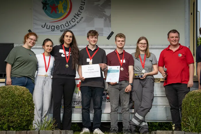 Sieben junge Menschen stehen auf einer Bühne vor einem LKW mit dem Aufdruck „Jugendrotkreuz Märkischer Kreis“ und halten Urkunden und Medaillen. Sie repräsentieren den Schulsanitätsdienst des Gertrud-Bäumer-Berufskollegs Lüdenscheid, der bei den Rotkreuzspielen den ersten Platz belegte.