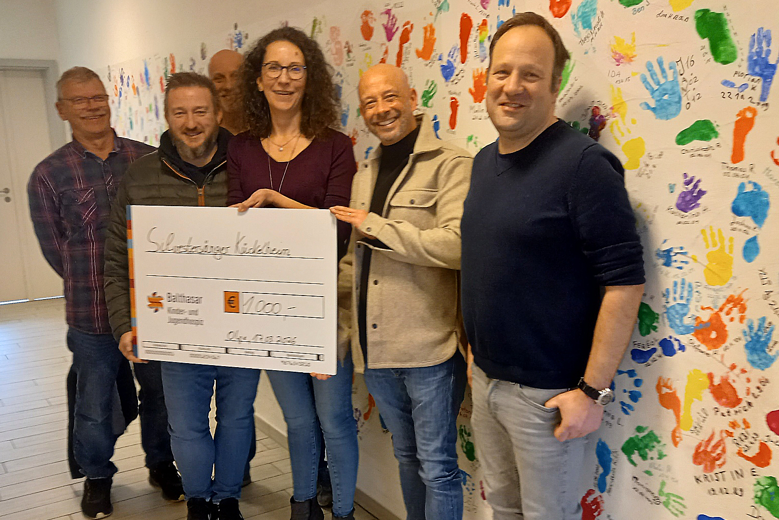 Fünf Männer und eine Frau überreichen einen Scheck in Höhe von 1000 Euro an Rebecca Kranz von Balthasar Kinder- und Jugendhospiz. Im Hintergrund eine Wand mit bunten Handabdrücken.