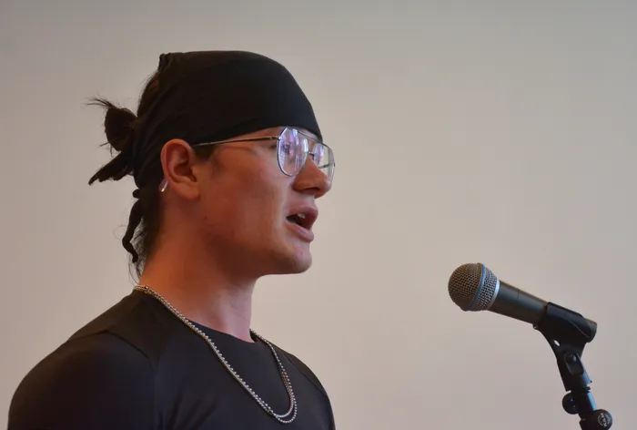Ein junger Mann mit schwarzem Bandana und Brille spricht während einer Performance in ein Mikrofon.