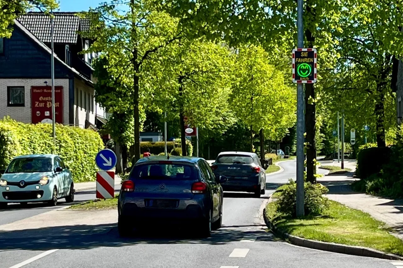 Autos fahren auf einer Straße mit Bäumen, an der ein Verkehrsschild mit einem Smiley-Gesicht angebracht ist, das die Geschwindigkeit anzeigt. Links steht ein Gebäude mit dem Schild