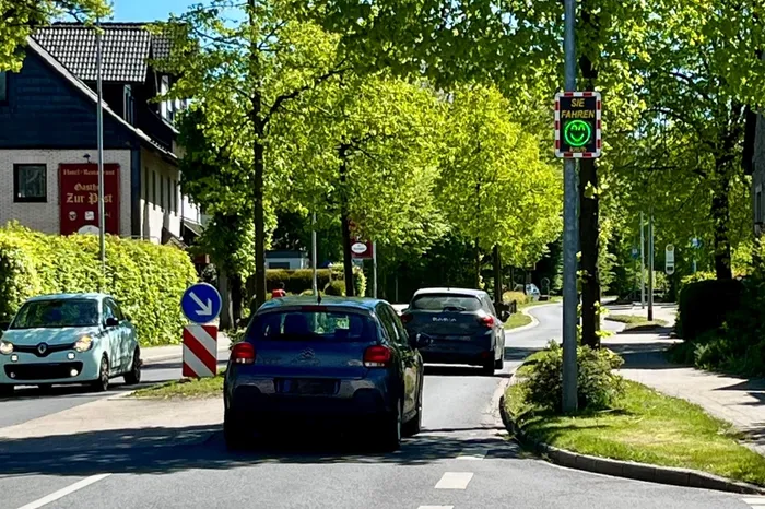 Autos fahren auf einer Straße mit Bäumen, an der ein Verkehrsschild mit einem Smiley-Gesicht angebracht ist, das die Geschwindigkeit anzeigt. Links steht ein Gebäude mit dem Schild