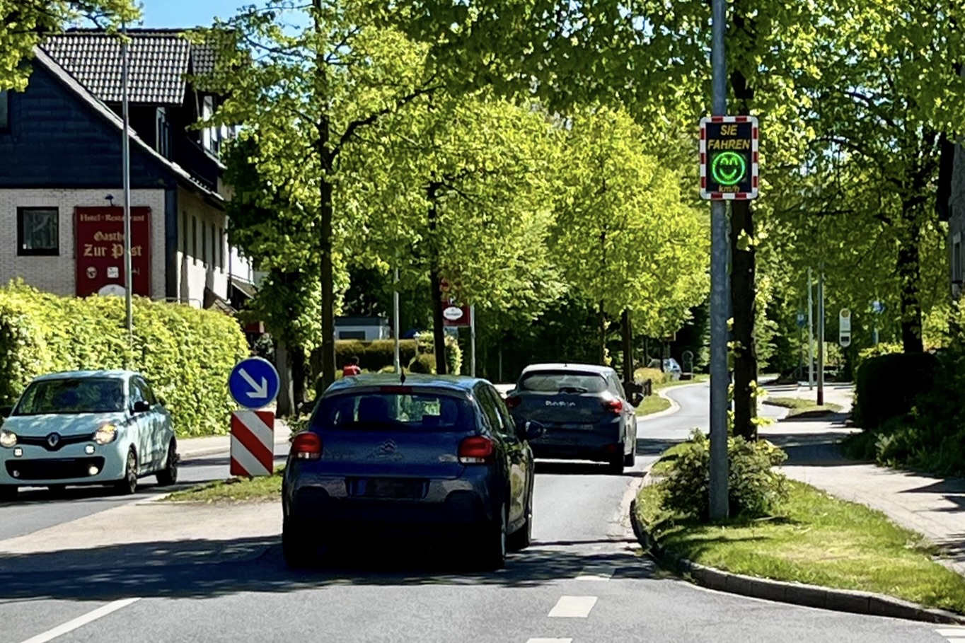 Autos fahren auf einer Straße mit Bäumen, an der ein Verkehrsschild mit einem Smiley-Gesicht angebracht ist, das die Geschwindigkeit anzeigt. Links steht ein Gebäude mit dem Schild