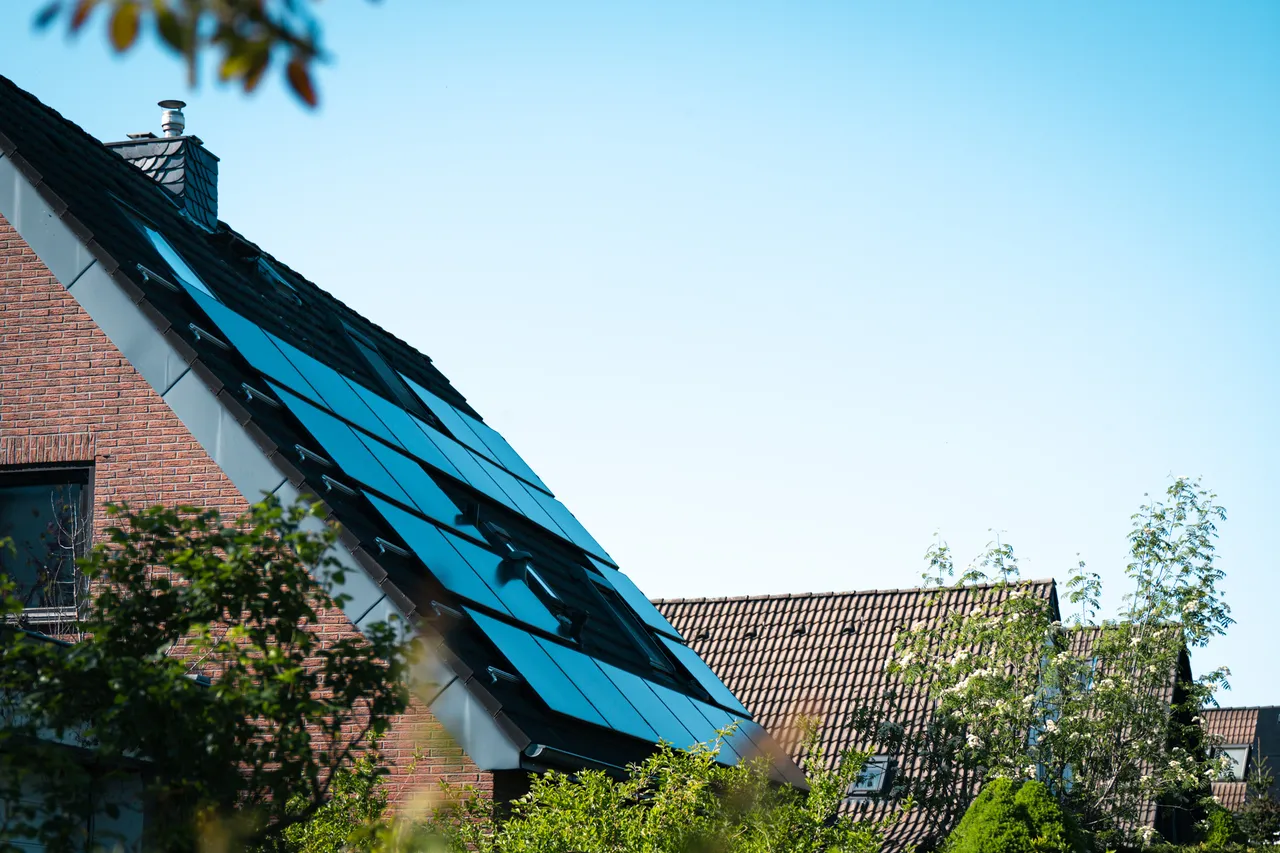 Solarpanels bedecken das Dach eines Ziegeldachhauses, umgeben von Grünflächen und unter blauem Himmel. Das Bild illustriert das Thema erneuerbare Energien und die Unterstützung durch die Verbraucherzentrale NRW.