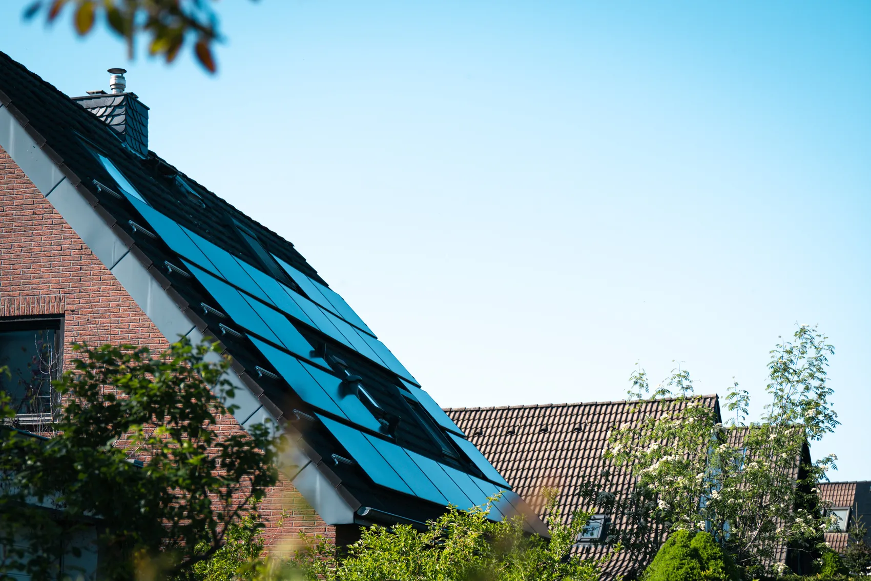 Solarpanels bedecken das Dach eines Ziegeldachhauses, umgeben von Grünflächen und unter blauem Himmel. Das Bild illustriert das Thema erneuerbare Energien und die Unterstützung durch die Verbraucherzentrale NRW.