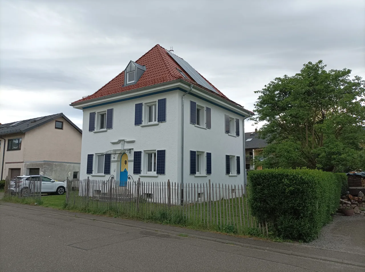 Ein zweistöckiges Haus mit einem roten Ziegeldach und blauen Fensterläden. Auf dem Dach sind Solarmodule installiert. Vor dem Haus befindet sich ein kleiner Vorgarten mit einem Holzzaun.