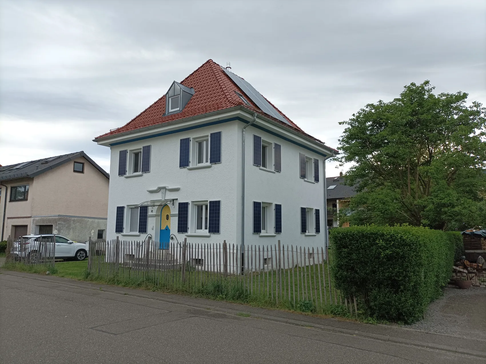 Ein zweistöckiges Haus mit einem roten Ziegeldach und blauen Fensterläden. Auf dem Dach sind Solarmodule installiert. Vor dem Haus befindet sich ein kleiner Vorgarten mit einem Holzzaun.