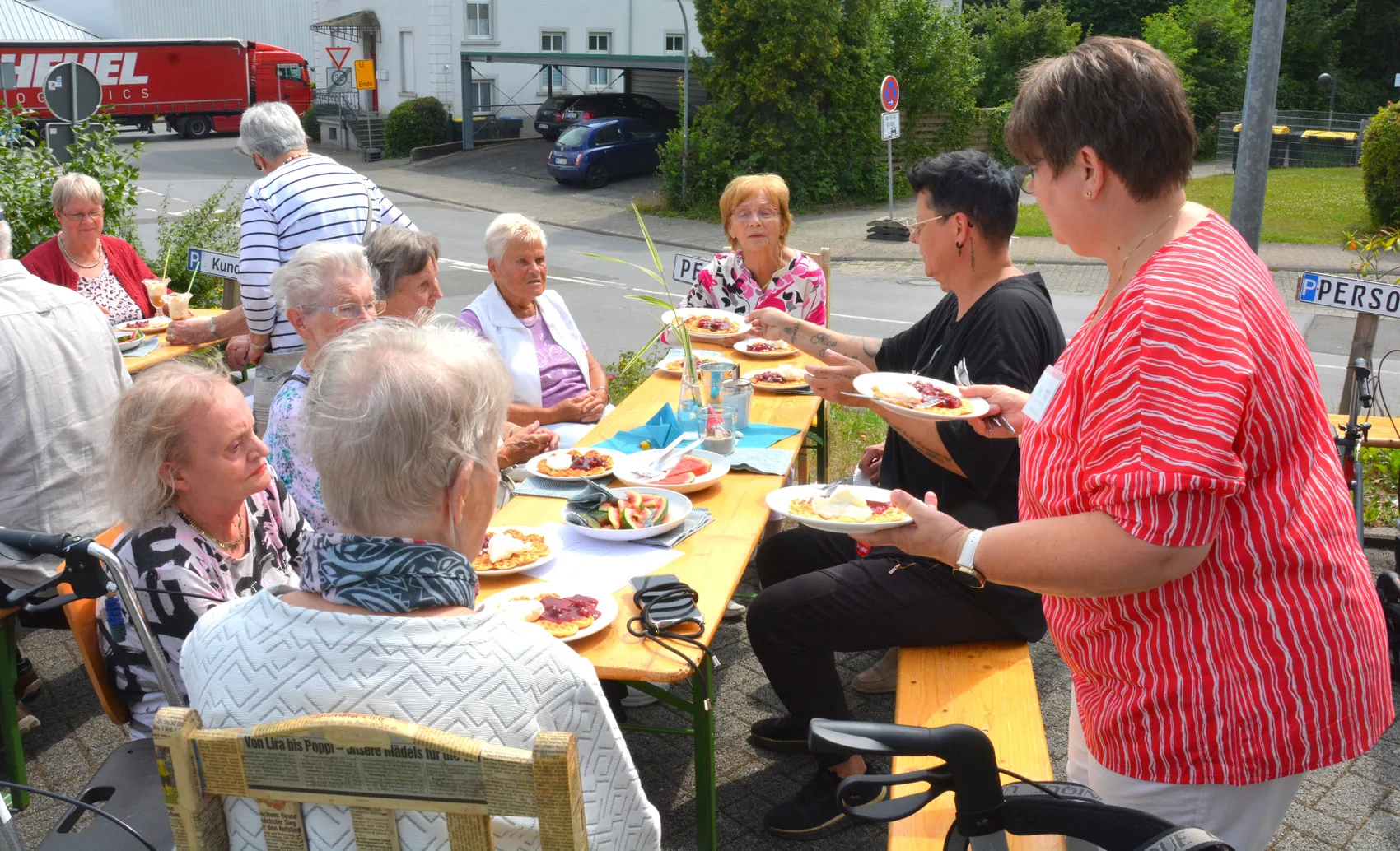 Eine Gruppe älterer Menschen sitzt an einem gedeckten Tisch im Freien und isst. Auf den Tellern sind verschiedene Speisen zu erkennen.