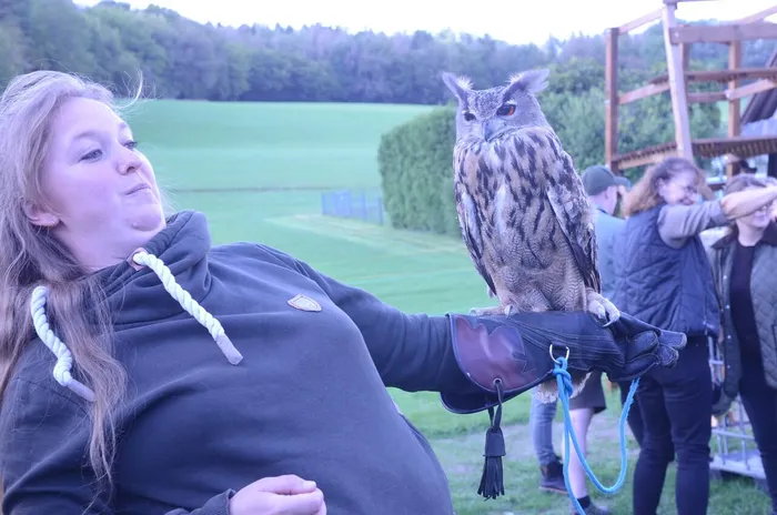 Eine Frau hält eine große Eule auf einem Lederhandschuh; im Hintergrund sind eine Wiese und weitere Personen zu sehen.
