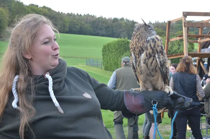 Eine Frau hält eine große Eule auf einem Lederhandschuh vor einer grünen Wiese und Bäumen.