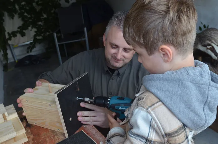 Ein Mann unterstützt einen Jungen beim Befestigen einer schwarzen Platte an einem Holzobjekt mit einem Akkuschrauber.