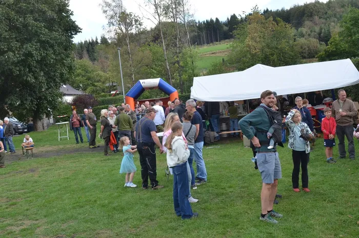 Menschen unterschiedlichen Alters feiern ein Sommerfest auf einer Wiese vor einem weißen Zelt und einem blauen aufblasbaren Torbogen.