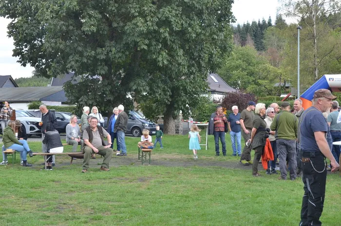 Eine Gruppe von Menschen verschiedener Altersgruppen versammelt sich bei einem Sommerfest auf einer Wiese vor einem großen Baum.