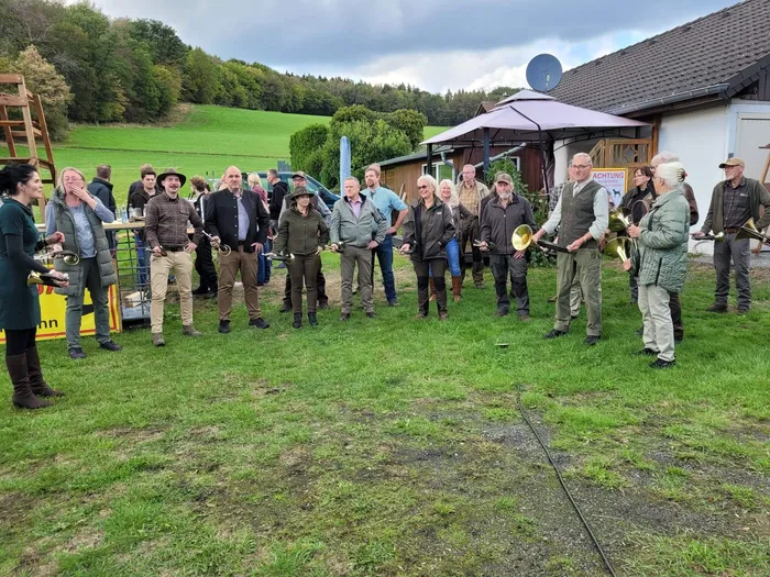 Eine Gruppe von Jägern und Jägerinnen steht auf einer Wiese vor einem Gebäude und einem Zelt; einige halten Blasinstrumente.