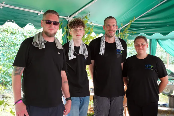 Vier Personen stehen unter einem grünen Zelt und schauen in die Kamera. Sie tragen schwarze T-Shirts, einige haben Tücher um den Nacken. Im Hintergrund sind Pflanzen und ein Garten zu sehen.