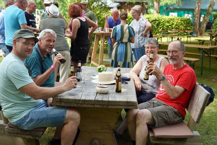 Vier Personen sitzen an einem Holztisch im Freien, Biergläser in der Hand, und unterhalten sich bei einem Sommerfest. Im Hintergrund sind weitere Menschen und Festzeltgarnituren zu sehen.