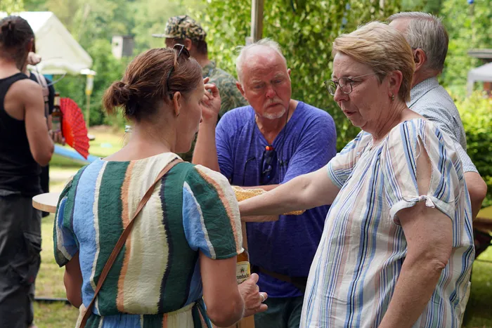 Zwei Frauen und ein Mann stehen im Freien und unterhalten sich. Die Frau rechts zeigt mit dem Finger auf die Maiskolben, die die Frau links hält. Im Hintergrund sind weitere Personen und Bäume zu sehen.