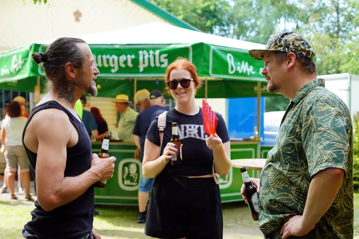Drei Personen stehen auf einer Wiese vor einem Getränkestand und unterhalten sich bei Bierflaschen in der Hand. Im Hintergrund sind weitere Menschen und ein grüner Stand mit der Aufschrift 
