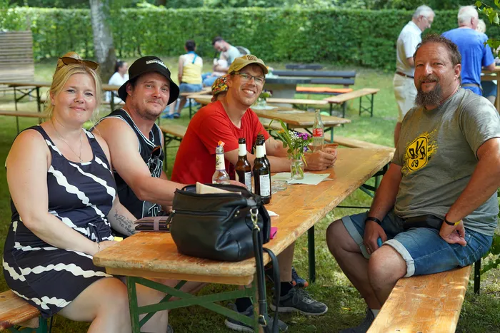Vier lächelnde Menschen sitzen an einem Holztisch im Freien, umgeben von weiteren Personen und Biergartenmöbeln. Auf dem Tisch stehen Getränke und eine kleine Blumenvasen.