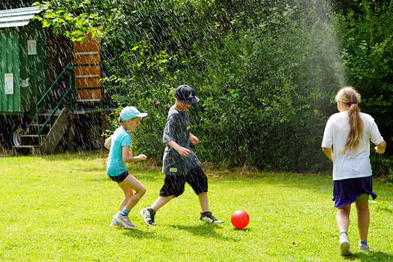 Drei Kinder spielen auf einer grünen Wiese unter einem Regenschauer mit einem roten Ball. Im Hintergrund steht dichtes Grün und ein Gebäude.
