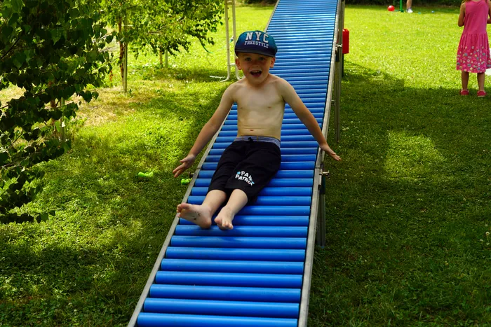 Ein Junge mit Baseballkappe rutscht lächelnd auf einer blauen Rollenbahn im Grünen herunter. Im Hintergrund sind weitere Personen und ein Garten zu sehen.