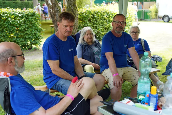 Vier Personen sitzen im Schatten auf einer Bank und unterhalten sich während eines Sommerfestes. Sie tragen blaue T-Shirts und wirken entspannt. Im Vordergrund steht ein Tisch mit Getränken und Sonnencreme.