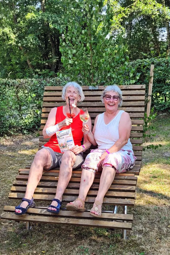 Zwei ältere Frauen sitzen lachend auf einer Holzbank im Freien, jede mit einem Glas in der Hand. Die Frau links hält zusätzlich eine Broschüre.