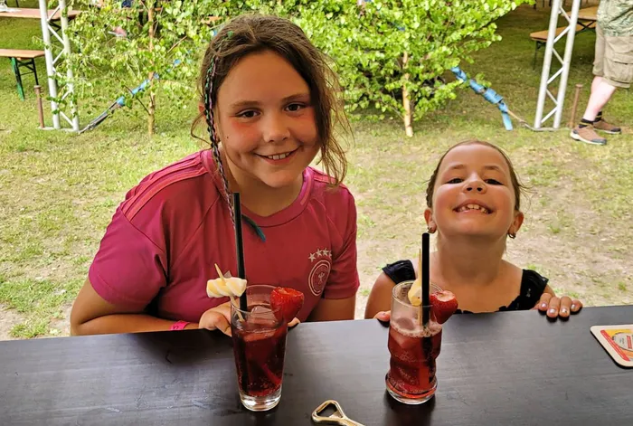 Zwei lächelnde Mädchen sitzen an einem Tisch und halten bunte Getränke mit Strohhalmen und Früchten. Im Hintergrund ist ein Festzelt und grüne Wiese zu sehen.