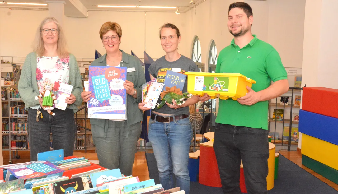 Vier Mitarbeitende der Stadtbücherei Lüdenscheid stehen mit Büchern und einer Kiste voller Spielzeug vor einem Bücherregal und stellen das Programm des Sommerleseclubs vor.