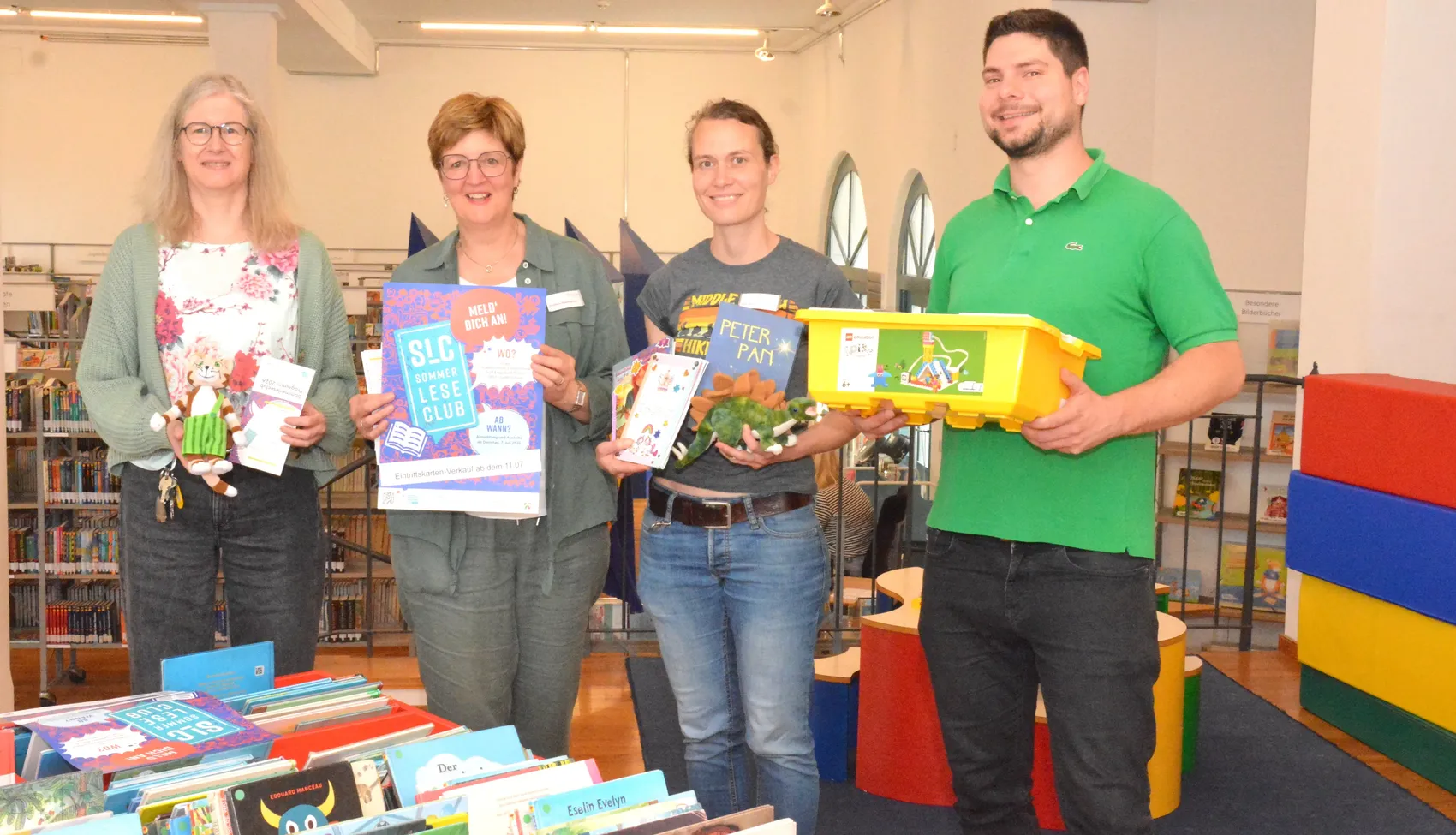 Vier Mitarbeitende der Stadtbücherei Lüdenscheid stehen mit Büchern und einer Kiste voller Spielzeug vor einem Bücherregal und stellen das Programm des Sommerleseclubs vor.