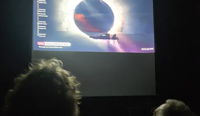 Projektion eines Livestreams einer totalen Sonnenfinsternis auf einer Leinwand vor einem Publikum in einem dunklen Raum.