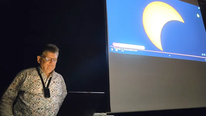 Rolf Becker steht an einem Laptop neben einer Leinwand, auf der eine Sonnenfinsternis projiziert wird.