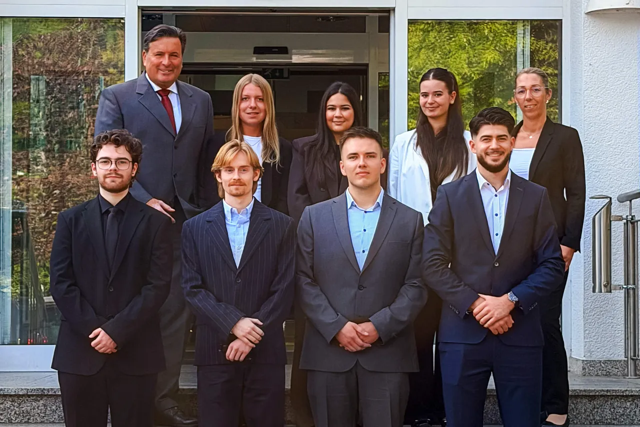Sieben Personen in Business-Kleidung stehen in zwei Reihen vor einem gläsernen Eingangsbereich eines Gebäudes.
