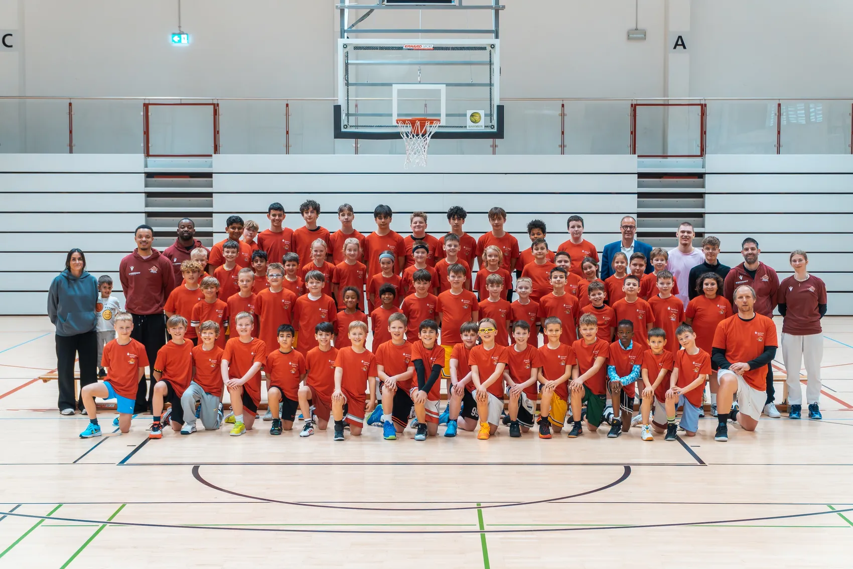 Gruppe von 66 Kindern und Jugendlichen in roten T-Shirts posiert in einer Basketballhalle vor einem Basketballkorb für ein Foto.