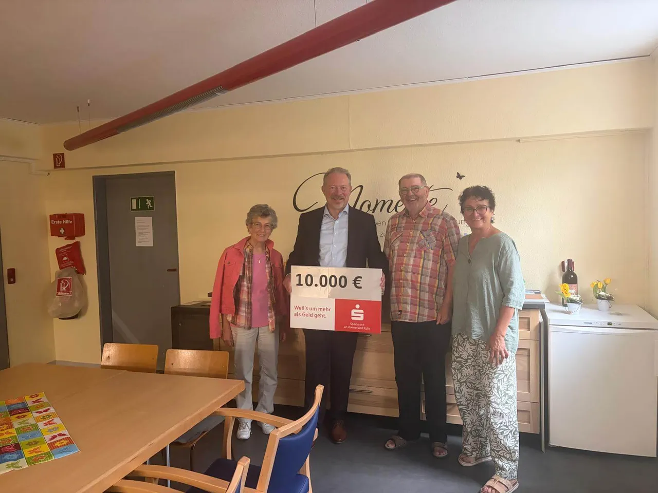 Vier Personen stehen in einem Raum mit einem übergroßen Scheck der Sparkasse an Volme und Ruhr über 10.000 Euro für die Quartiersarbeit der Arche.
