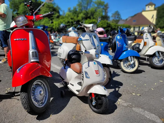 Rote und weiße Vespas stehen nebeneinander auf einem Parkplatz, im Hintergrund weitere Roller und Bäume. Die Aufnahme zeigt eine Spendentour des Vespa-Clubs Plettenberg für krebskranke Kinder.