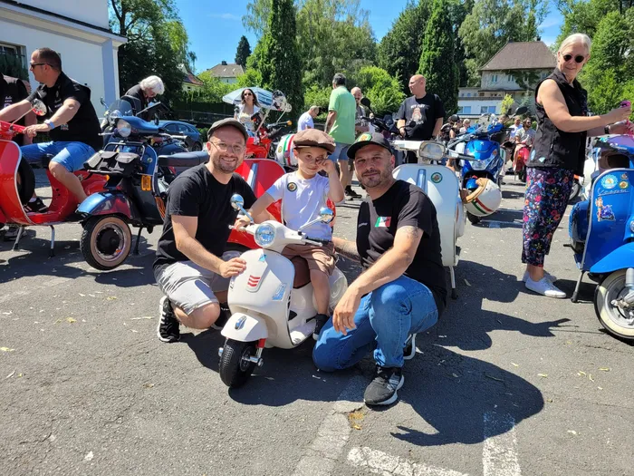 Drei Personen lächeln vor einer hellblauen Vespa, im Hintergrund weitere Menschen und Vespas während einer Spendentour für krebskranke Kinder.