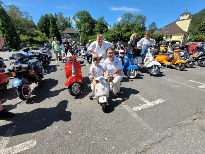 Ein Mann, eine Frau und ein Kind sitzen auf roten und weißen Vespas vor einer Gruppe weiterer Vespas und Menschen im Freien. Im Hintergrund sind Gebäude und Bäume zu sehen. Die Aufnahme entstand bei der Spendentour des Vespa-Clubs Plettenberg für krebskranke Kinder.