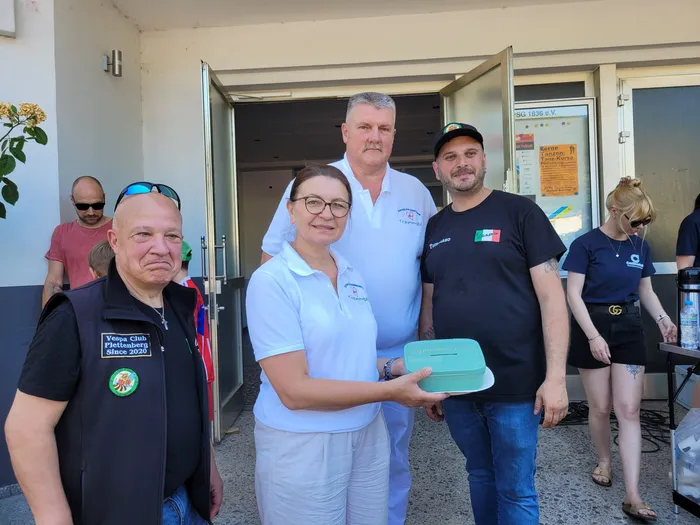 Vier Personen überreichen eine Spendenbox vor dem Eingang eines Gebäudes. Ein Mann trägt eine Weste mit dem Aufdruck 