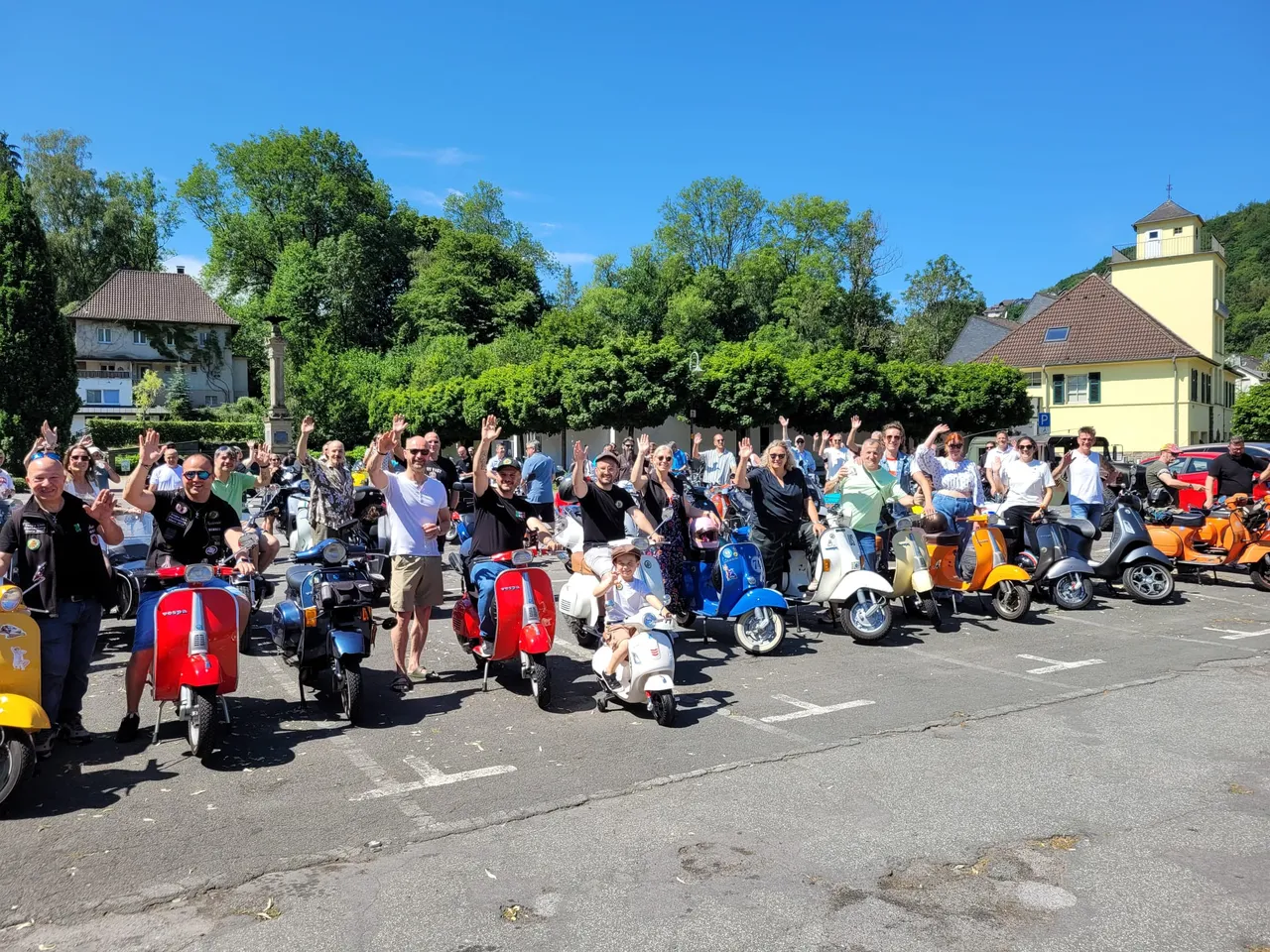 Eine Gruppe von Menschen steht vor ihren Vespas und winkt in die Kamera. Im Hintergrund sind Häuser und Bäume zu sehen.
