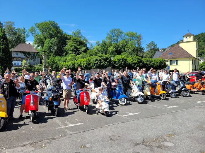 Eine Gruppe von Menschen steht vor ihren Vespas und winkt in die Kamera. Im Hintergrund sind Häuser und Bäume zu sehen.
