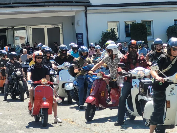 Eine Gruppe von Menschen auf Vespas nimmt an einer Wohltätigkeitsfahrt teil. Die Fahrer tragen Helme und fahren auf einer Straße vor hellen Gebäuden.