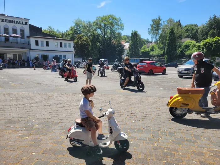 Ein Junge mit einer braunen Schirmmütze sitzt auf einer weißen Vespa. Im Hintergrund stehen mehrere Menschen und weitere Vespas auf einem Platz vor einem hellen Gebäude.