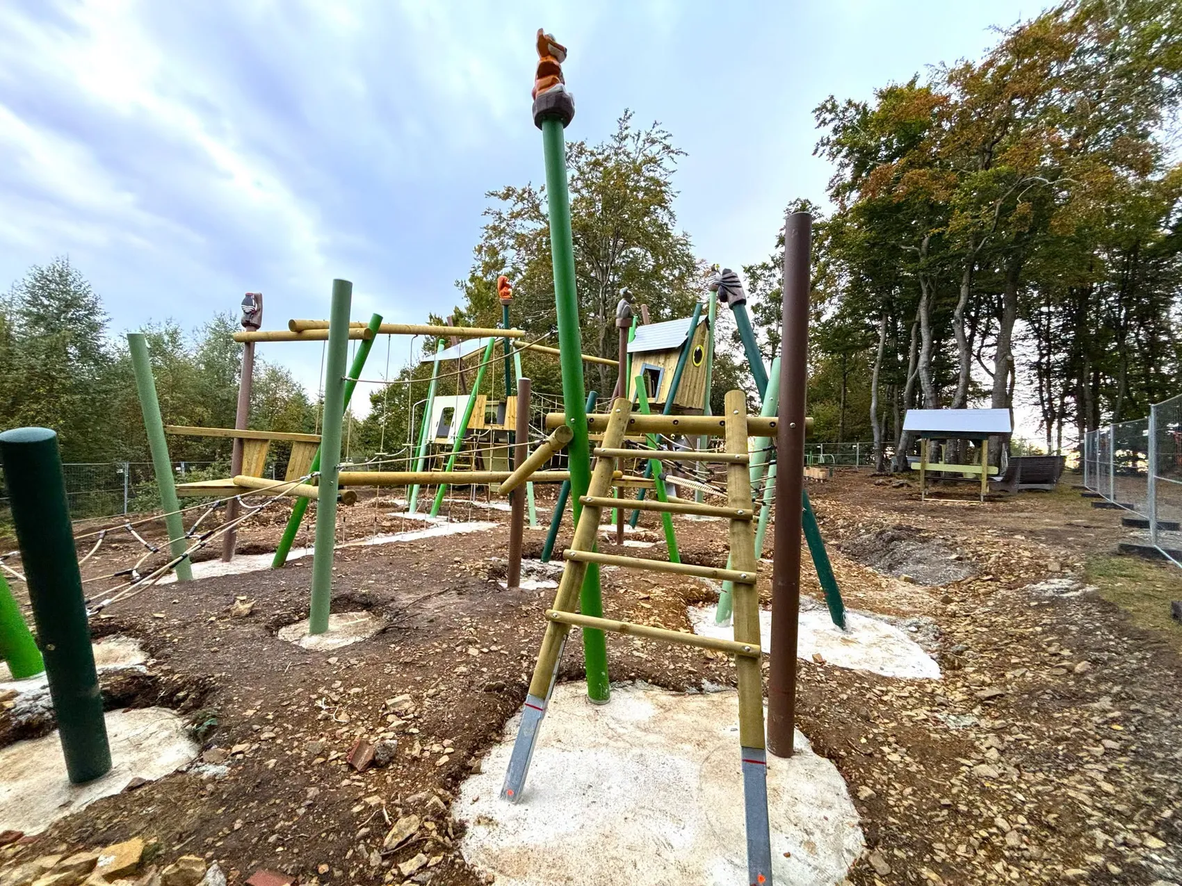Verschiedene Klettergerüste aus Holz und Seilen auf einem im Bau befindlichen Spielplatz mit Bäumen im Hintergrund.