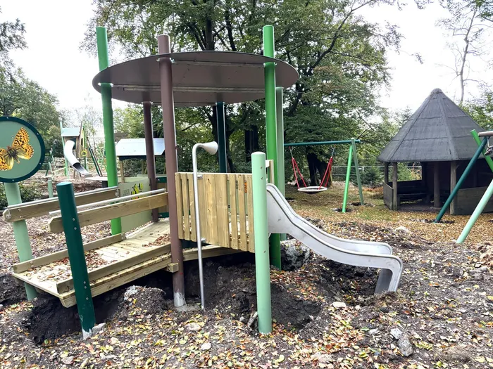 Ein hölzernes Spielgerät mit einer weißen Rutsche und einem Dach steht auf einem mit Laub bedeckten Erdhügel.