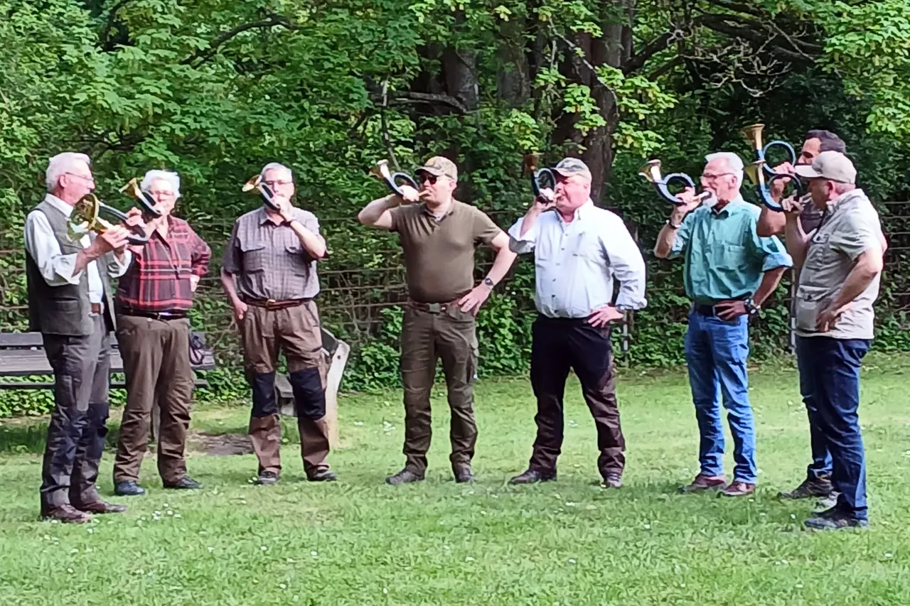 Eine Bläsergruppe spielt auf einer grünen Wiese vor Bäumen. Die Musiker, allesamt Männer mittleren Alters, spielen verschiedene Blechblasinstrumente.