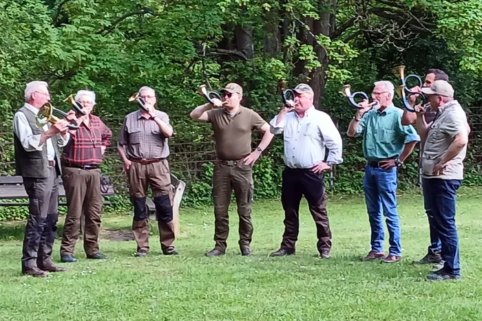 Eine Bläsergruppe spielt auf einer grünen Wiese vor Bäumen. Die Musiker, allesamt Männer mittleren Alters, spielen verschiedene Blechblasinstrumente.