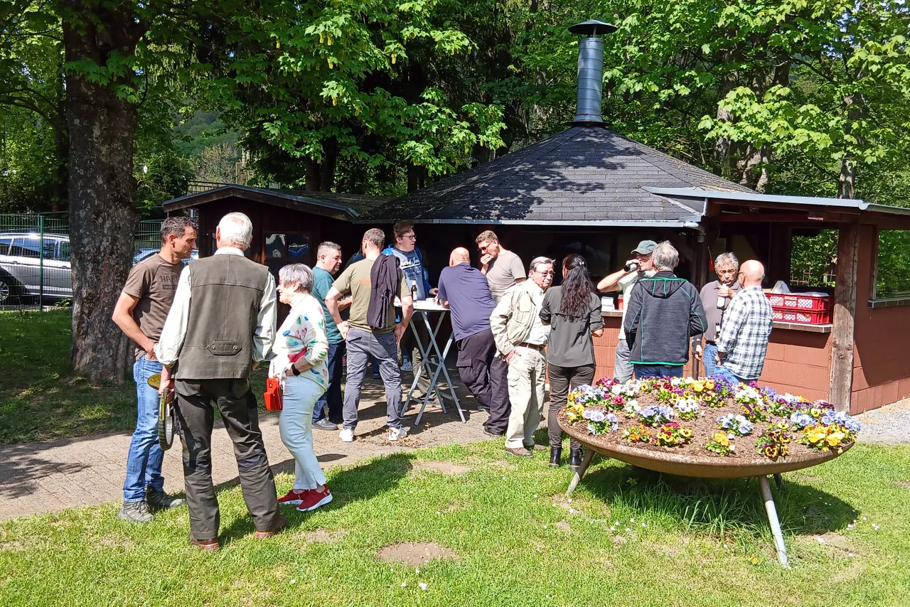 Menschen stehen vor einem Grillstand im Freien, einige unterhalten sich, andere warten auf Essen. Im Hintergrund Bäume und ein Auto.